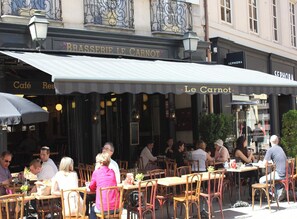 En udendørs café med borde, stole og et markise, samt en butiksfacade med skilte.