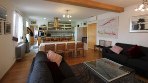 Ein modernes Wohnzimmer mit einer Couch, einem Glastisch und einem Essbereich, im Hintergrund ist eine Küche erkennbar.