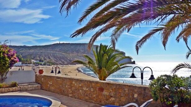Zona de piscina con sillones de descanso, vista a la playa y montañas al fondo.