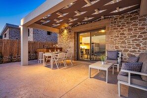 Un patio exterior con paredes de piedra, una pérgola de madera y una zona de comedor con mobiliario blanco.