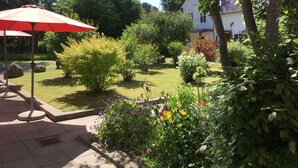 Een tuin met een rode parasol, verschillende planten en een wit gebouw op de achtergrond.