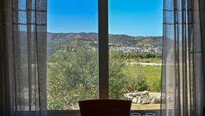 A room with a large window overlooking a mountainous landscape with greenery and a small settlement. - Olive Garden house (Καμηλάρι)