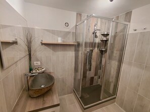 Ett modernt badrum med en duschavdelning med glasväggar, ett badrumsskåp i trä med en skålformad tvättställ och en liten hylla med toalettartiklar.