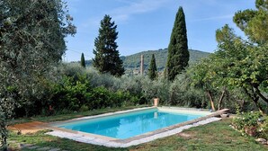 A rectangular swimming pool surrounded by a grassy area with stone edging, trees, and a hilly landscape in the background. - CASALE ASCIOLO (Le Case San Romolo)