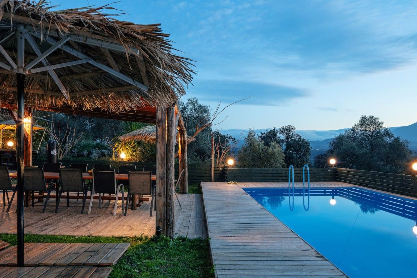 Une terrasse en bois avec une piscine, un espace salon extérieur et une structure surmontée d’un toit de chaume.