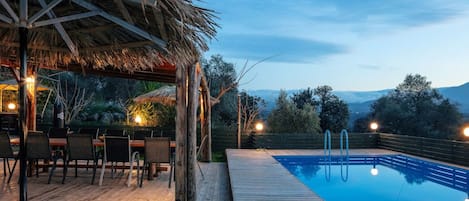 Une terrasse en bois avec une piscine, un espace salon extérieur et une structure surmontée d’un toit de chaume.