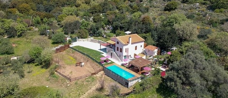 Une maison à deux étages avec une piscine, entourée d’une végétation luxuriante et de collines.