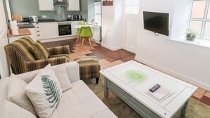 A living room with a sofa, a coffee table, and a dining area with chairs and a table. There is a kitchen in the background with cabinets and appliances.