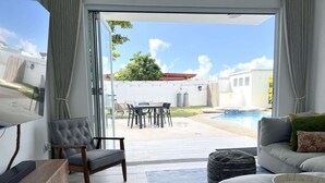 A modern living room with a sofa, a chair, and a TV on a wooden cabinet. There is a sliding glass door leading to a patio with a pool and outdoor furniture. - Luxury 5 Bedroom Home, 3 Minute Walk from the Beach (Dorado)
