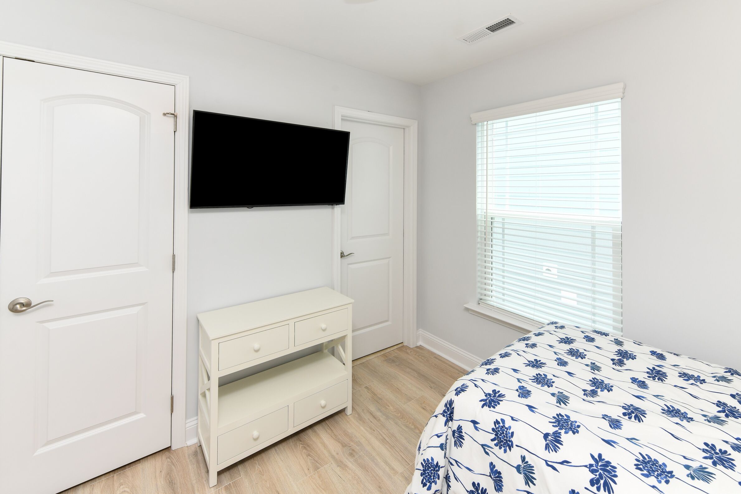Ein Schlafzimmer mit Bett, Wandfernseher, Kommode und Fenster mit Jalousien.