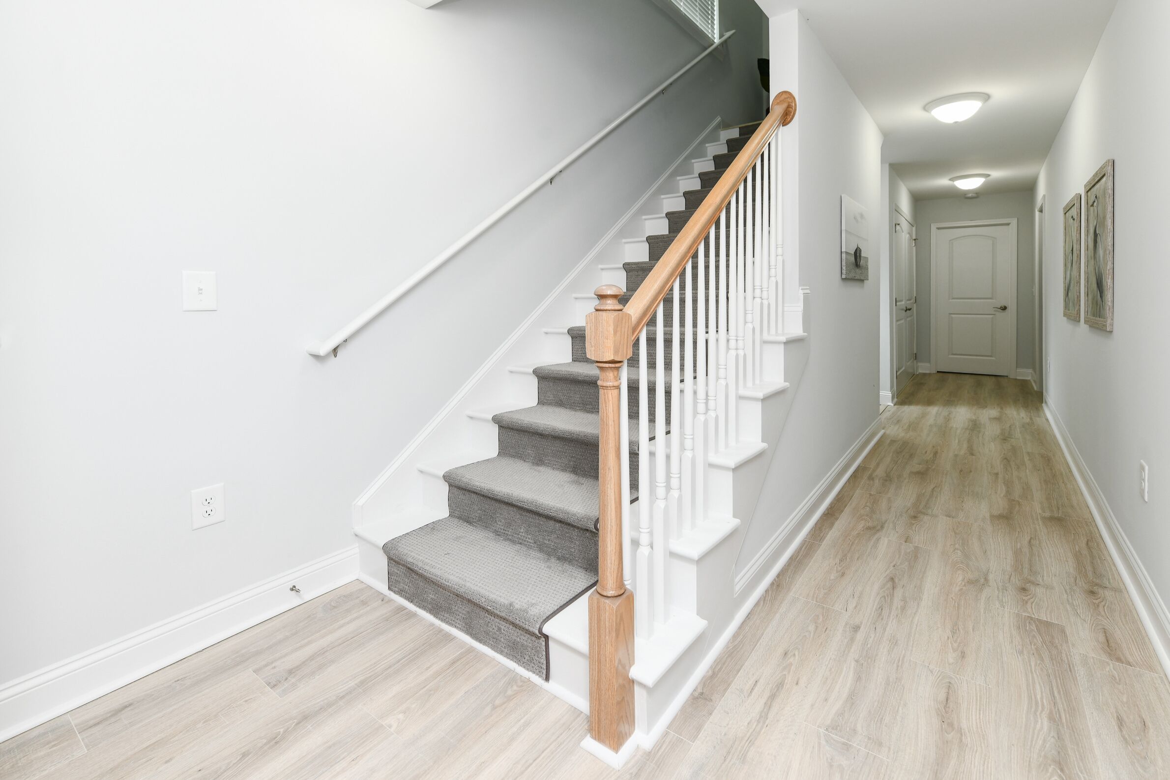 A staircase with wooden handrails and carpeted steps leading to a door at the end of a hallway with light wooden flooring.