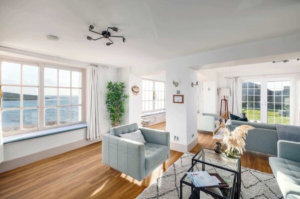 A spacious living room with large windows, a grey sofa, a coffee table, and a view of the sea.