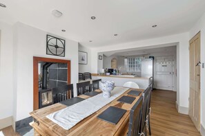 A dining area with a wooden table, chairs, and a white table runner. A kitchen is visible in the background with a refrigerator and a stove.