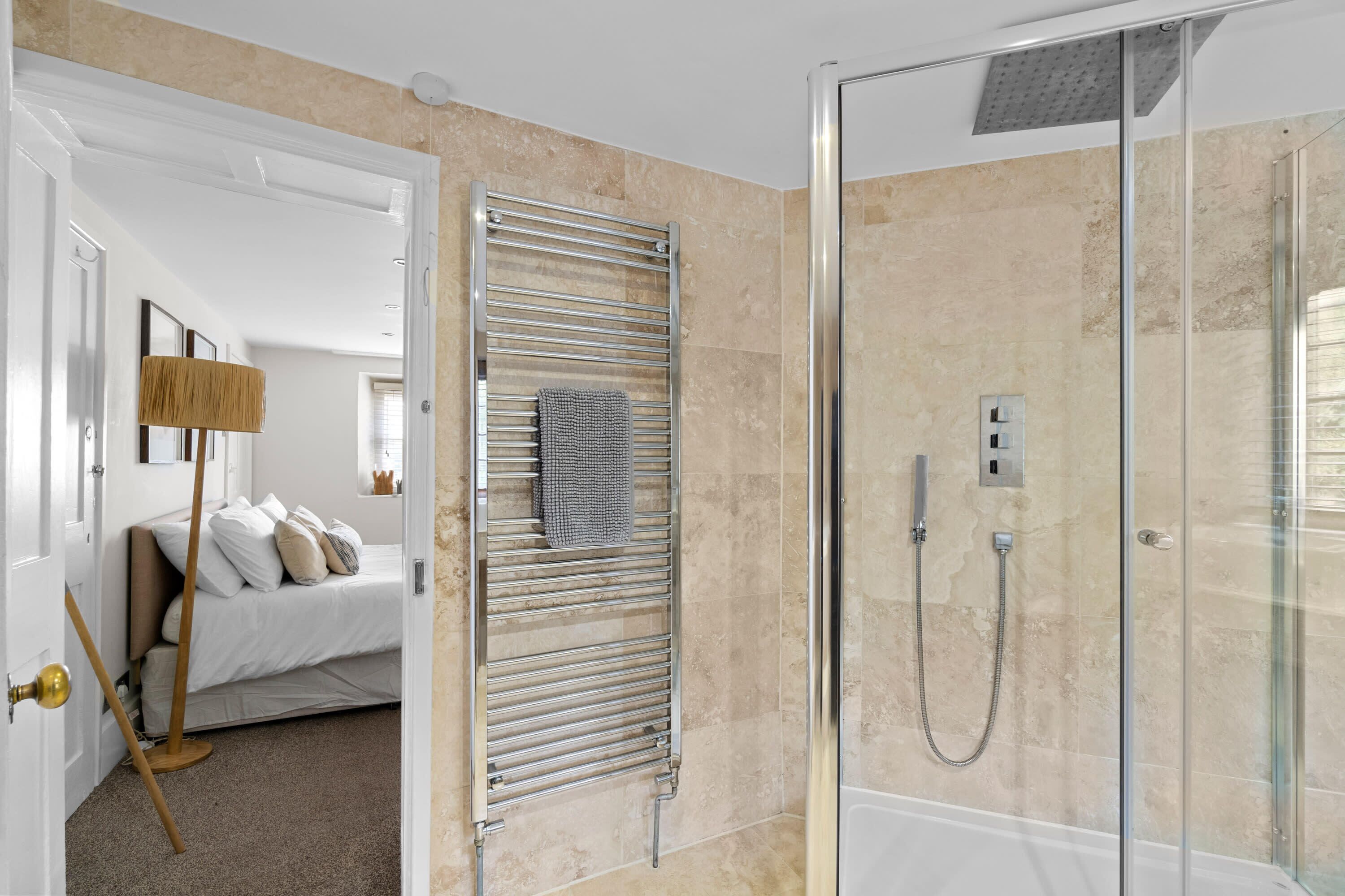 A modern bathroom with a glass shower enclosure, a towel rack, and a bed in the adjacent room.