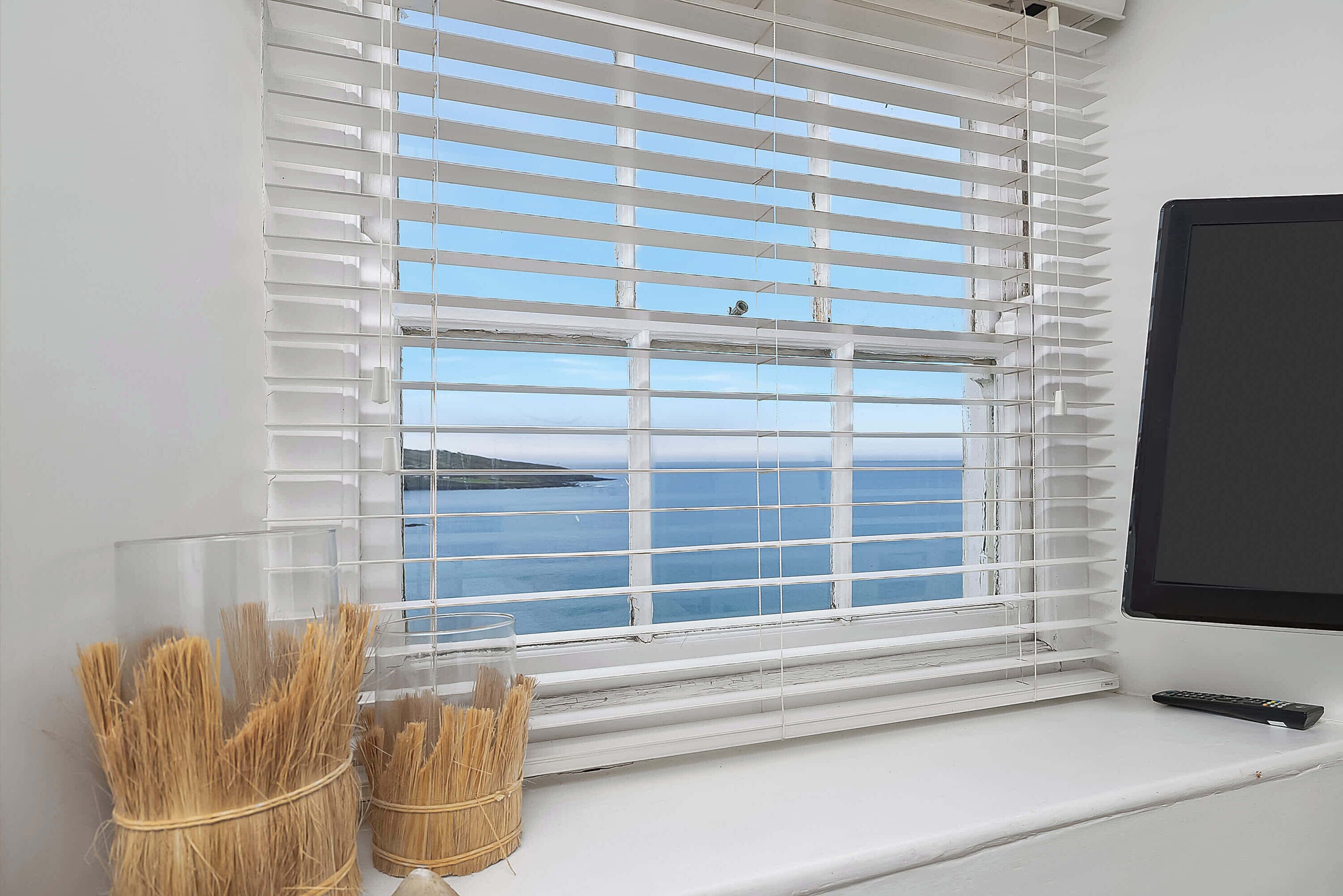 Una stanza con una finestra vista mare, una TV sulla destra e un piccolo ripiano con un contenitore di vetro e piante essiccate.