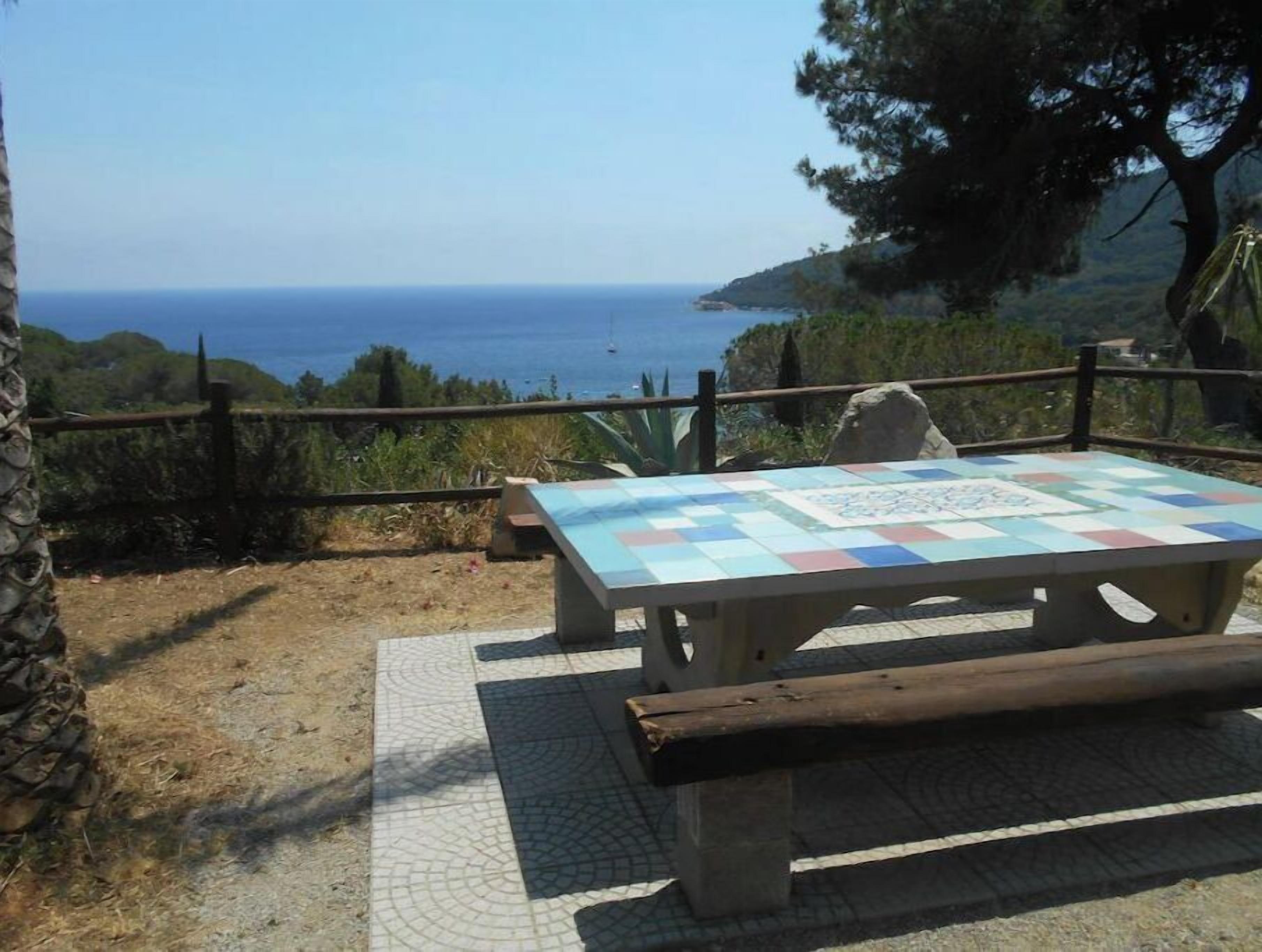 A picnic table with a colorful quilted tablecloth, positioned on a tiled patio with a view of the sea.