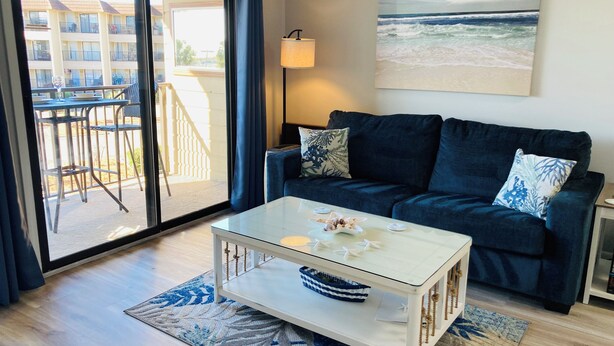 A living room with a glass coffee table, a dark blue sofa, a view of a building through a glass door, and a beach scene painting on the wall. - Ocean View 1-Bedroom. Steps to the beach. Sleeps 6. (Hilton Head Island)