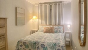 A bedroom with a bed, a nightstand, a dresser, a mirror, and a wall-mounted lamp. - Ocean View 1-Bedroom. Steps to the beach. Sleeps 6. (Hilton Head Island)