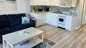 A compact kitchen with white cabinets, a microwave, a stove, and a dishwasher. There's a small dining area with a table and chairs, a couch, and a rug. - Ocean View 1-Bedroom. Steps to the beach. Sleeps 6. (Hilton Head Island)