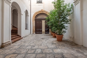 A narrow stone-paved courtyard with a staircase on the left, a large wooden door in the center, and potted plants on the right.