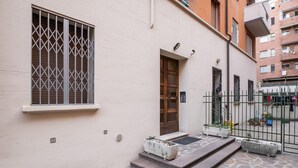 A building with a white facade, a barred window, a wooden door, and a small porch with potted plants. - Porta Mascarella Apartment by Wonderful Italy (Bologna)