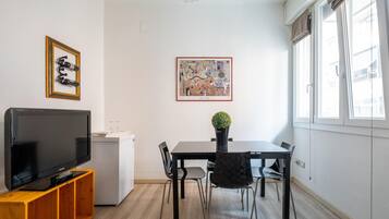 Una sala moderna con televisor de pantalla plana sobre un mueble de madera, una mesa de comedor con sillas, un refrigerador pequeño y una obra de arte enmarcada en la pared.