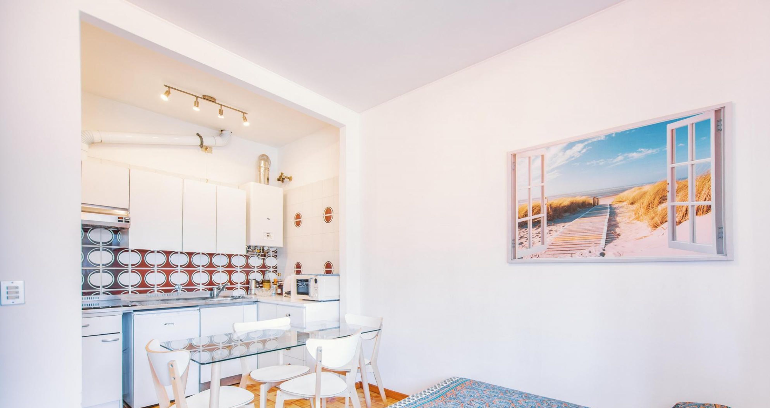 A compact kitchen with white cabinets, a glass dining table, and a beach scene painting.