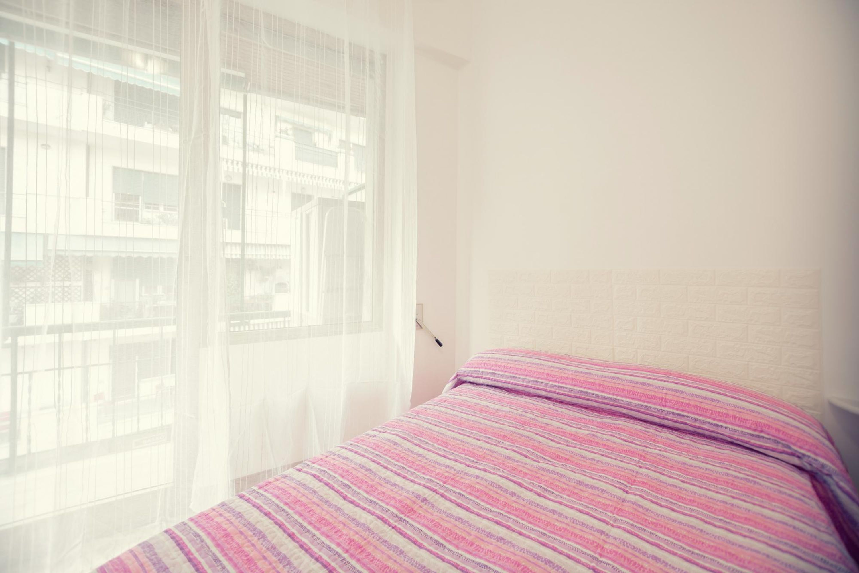 A bedroom with a bed, a window with sheer curtains, and a white brick wall.