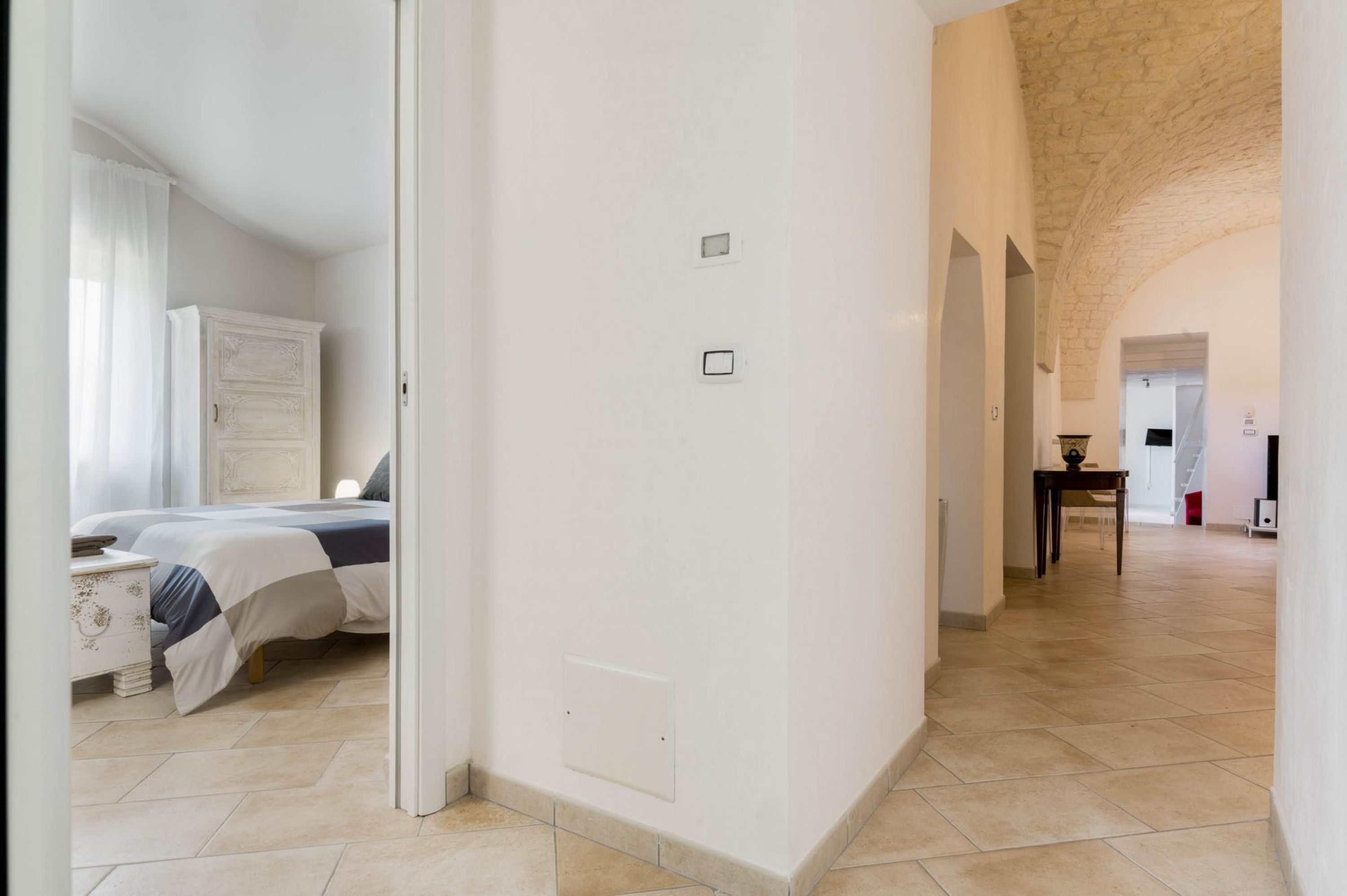 A spacious room with a bed, a nightstand, and a wardrobe. There is a hallway leading to another room.