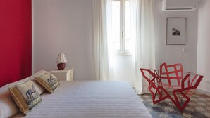 Une chambre à coucher avec un lit, une table de chevet, une lampe, une fenêtre avec des rideaux et un fauteuil rouge.