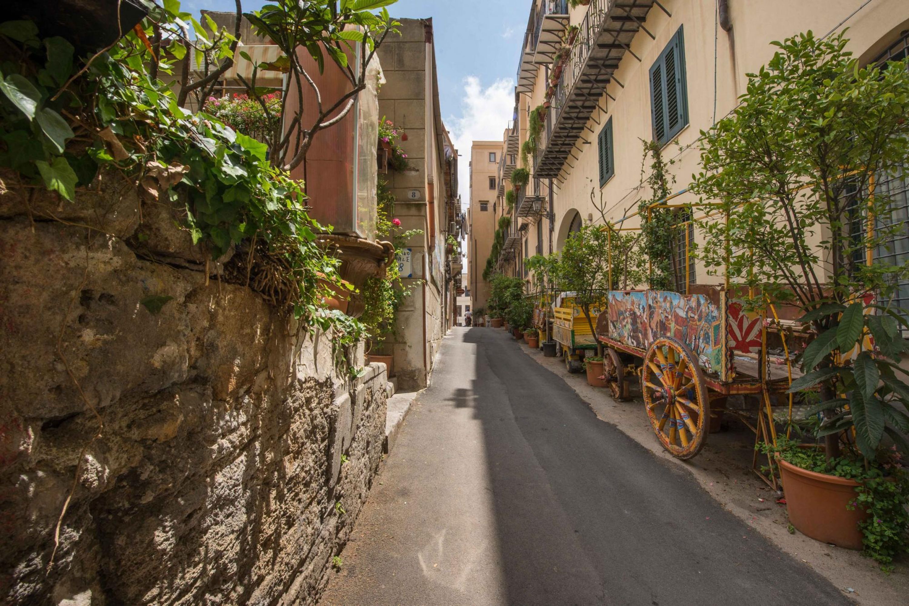 A narrow street with buildings on either side, a stone wall on the left, and a colorful cart on the right.