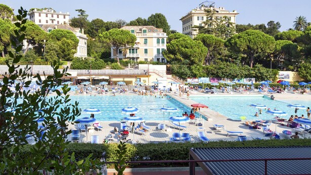 A large outdoor swimming pool surrounded by lounge chairs and umbrellas, with buildings and trees in the background. - Casa Rosanna by Wonderful Italy (Genoa)