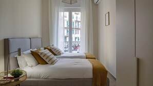 A bedroom with two beds, a bedside table, a lamp, and a window with a view of buildings. - Carignano Design Apartment 7 by Wonderful Italy (Genoa)