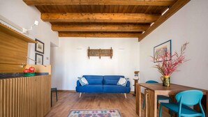 Een knus woongedeelte met een blauwe bank, houten balken in het plafond, een houten eettafel met stoelen en een vloerkleed met een patroon.