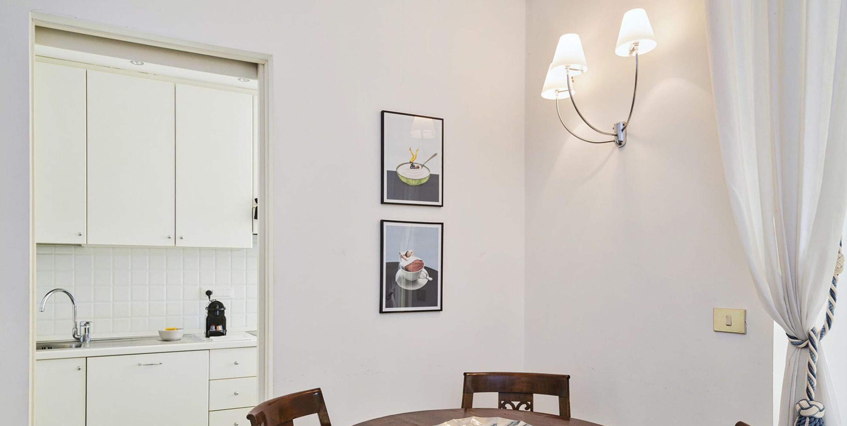 Een keuken met witte kastjes, een spoelbak en een koffieapparaat. Twee ingelijste foto's hangen aan de muur en er is een wandlamp.