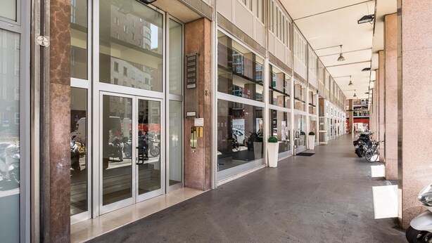 A corridor with glass storefronts, a tiled floor, and a row of parked scooters. - Cineteca White Studio by Wonderful Italy (Bologna)