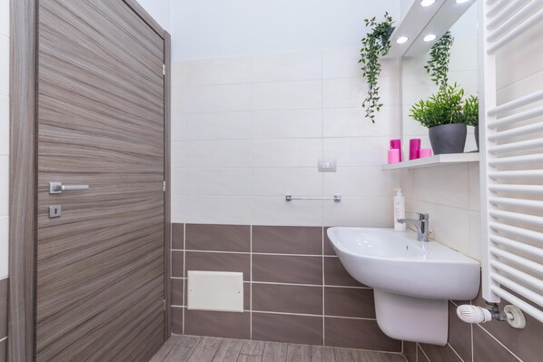 A modern bathroom with a wooden door, white tiles, a white sink, and a towel rack with plants. - Housing Giulia - Monolocale 5 by Wonderful Italy (Turin)