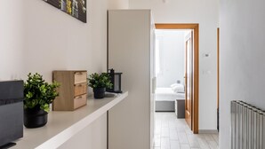 Un pasillo con un estante blanco que contiene plantas en macetas y un baúl de madera, que conduce a un dormitorio con cama y mesita de noche.