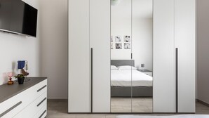 Un dormitorio moderno con un armario grande, un televisor de pantalla plana, una mesita de noche con lámpara y una cama con ropa de cama blanca.