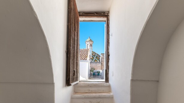 Eine schmale Treppe führt zu einer offenen Tür, von der aus man ein Gebäude mit einem Ziegeldach und einem Turm sieht.