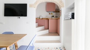 A compact living space with a TV mounted on the wall, a wooden dining table, a blue chair, a kitchen area with pink cabinets, and a white refrigerator. - Olivia in Wonderland by Wonderful Italy (Ostuni)