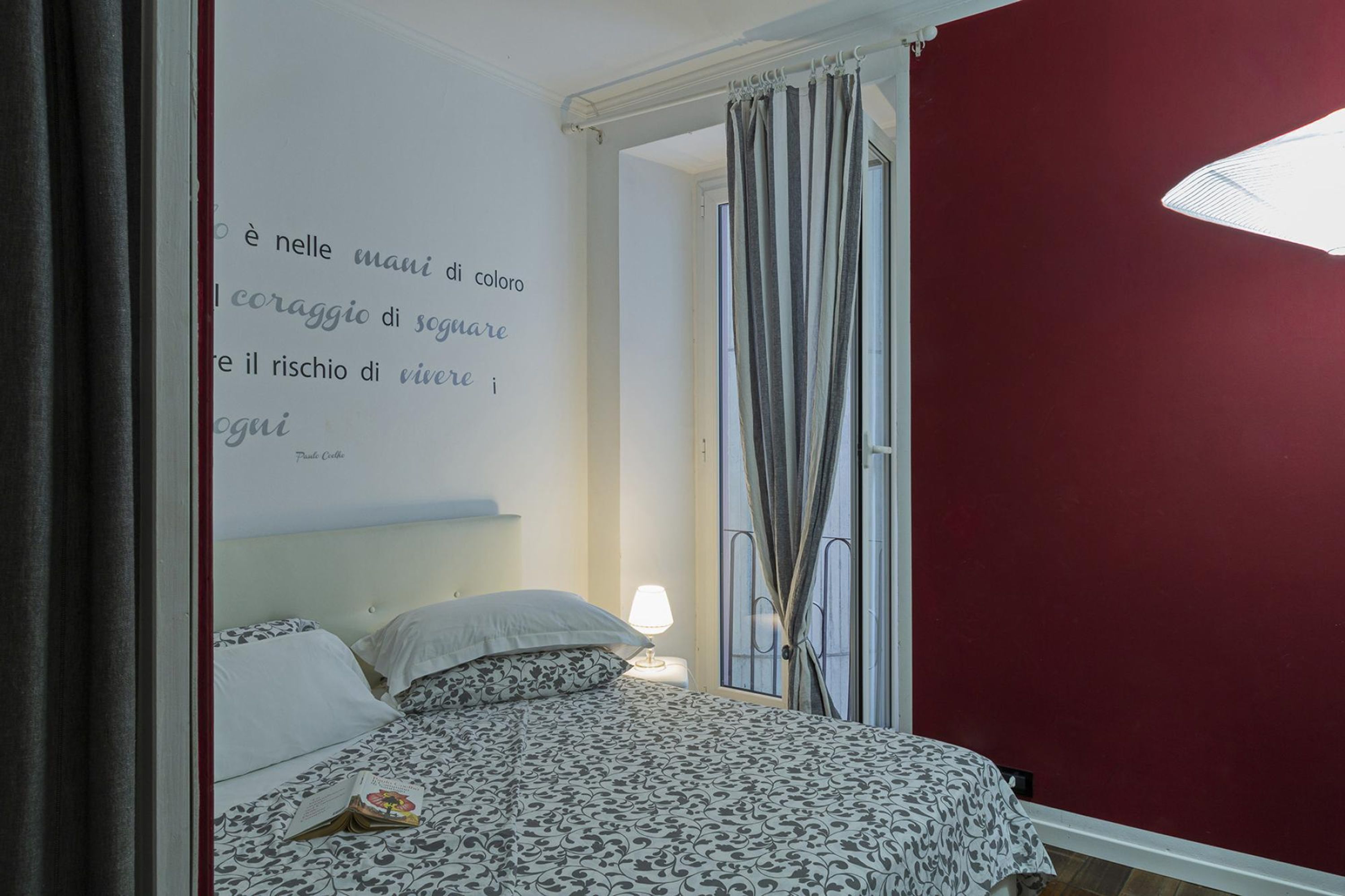 Un dormitorio con cama, mesita de noche, lámpara, ventana con cortinas y una pared con una cita.