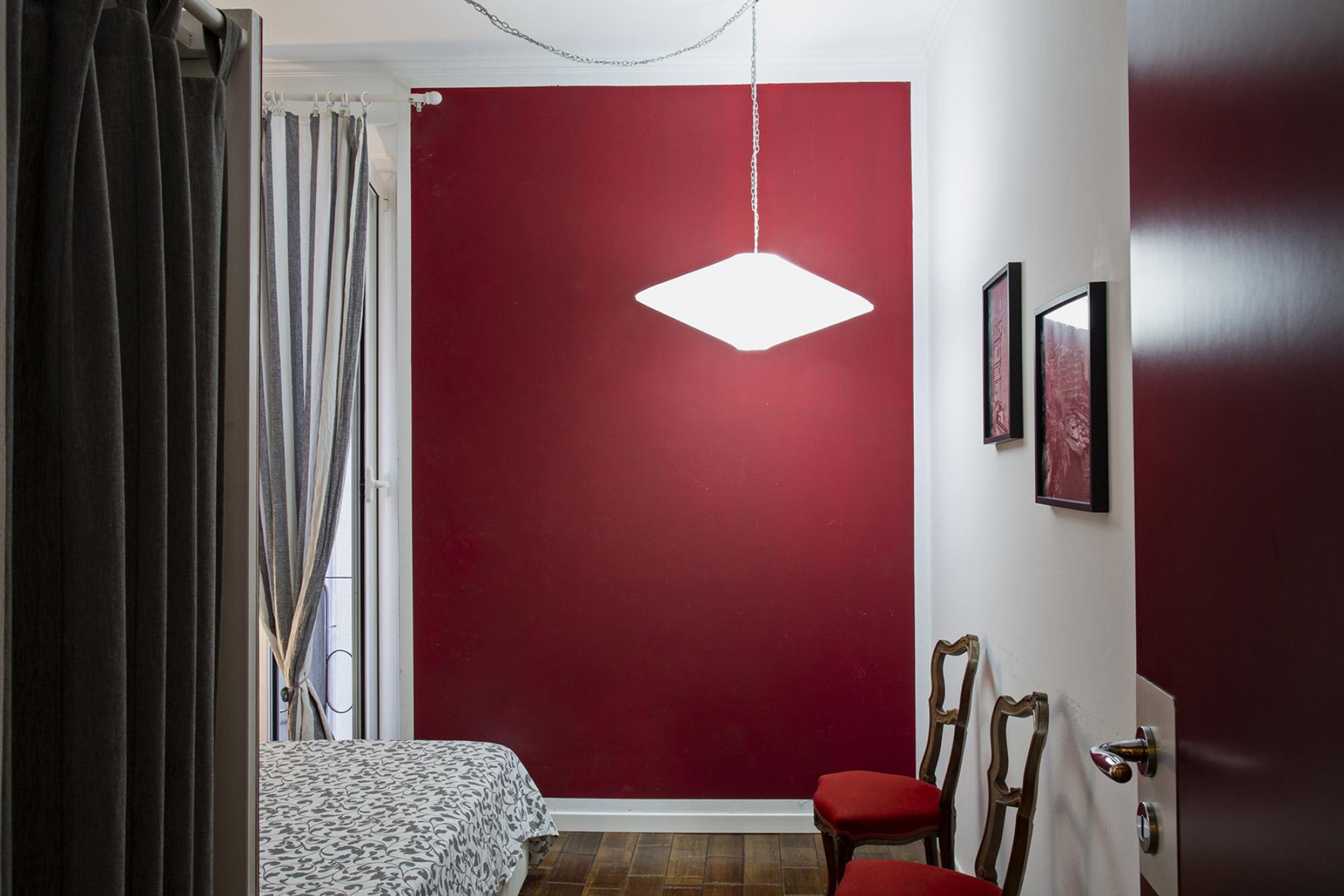 A bedroom with a red accent wall, a hanging light fixture, a bed with patterned bedding, and a chair.