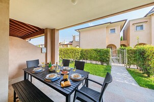 An outdoor dining area with a table set for four, surrounded by chairs, with a view of a residential building and a well-maintained hedge in the background.