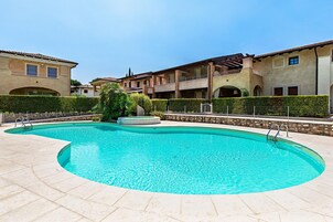 A swimming pool with a curved edge, surrounded by a paved area and a hedge.