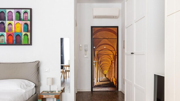 Habitación de hotel con cama, mesita de noche con lámpara, televisor, una obra de arte colorida en la pared y un pasillo que conduce a una escalera.