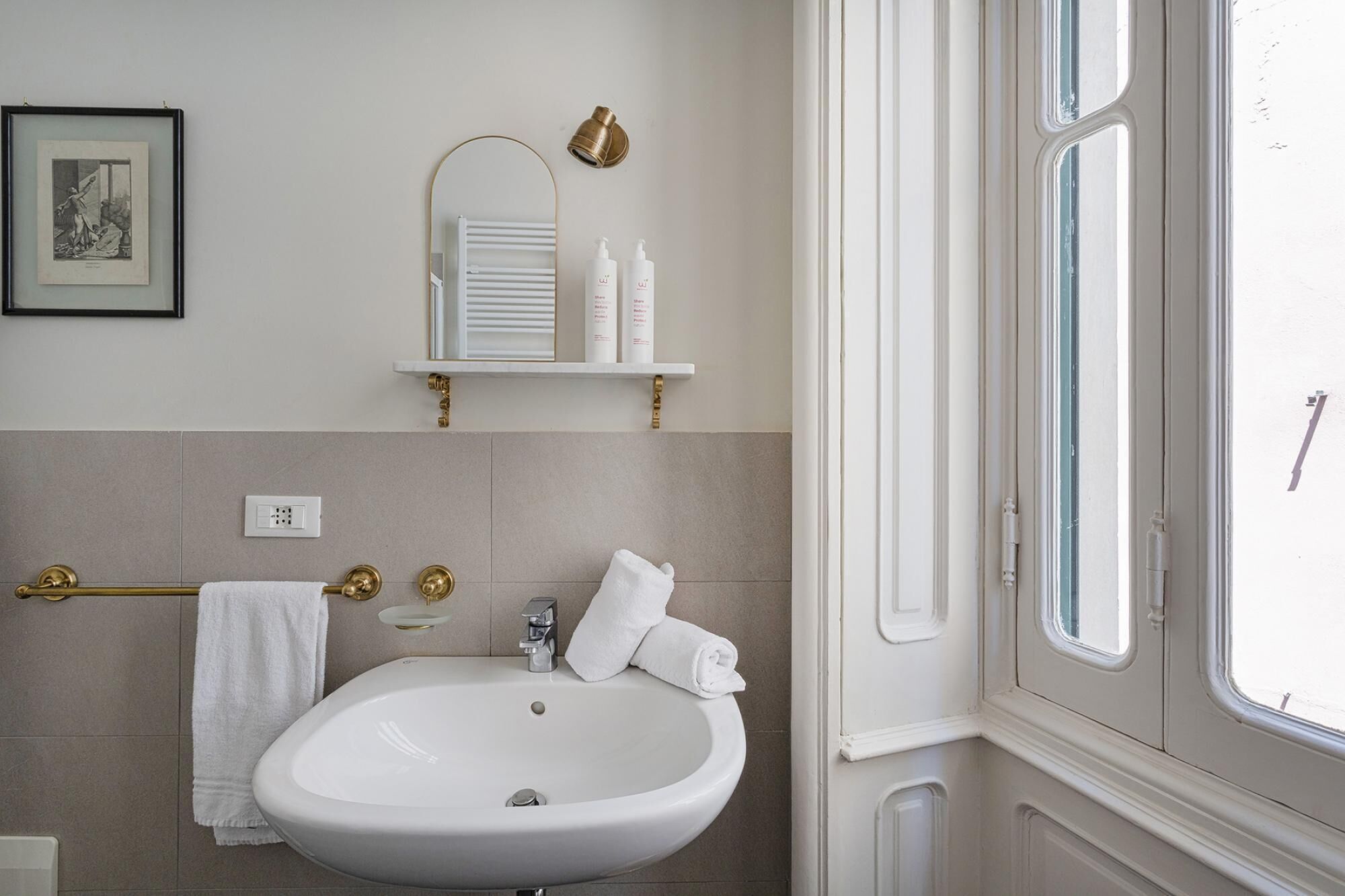 A bathroom with a white sink, a towel rack, a framed picture, a mirror, and a window.