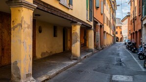A narrow street with yellow buildings, some with green shutters. There are parked scooters on the right. - Il Giardino Segreto di Orfeo by Wonderful Italy (Bologna)