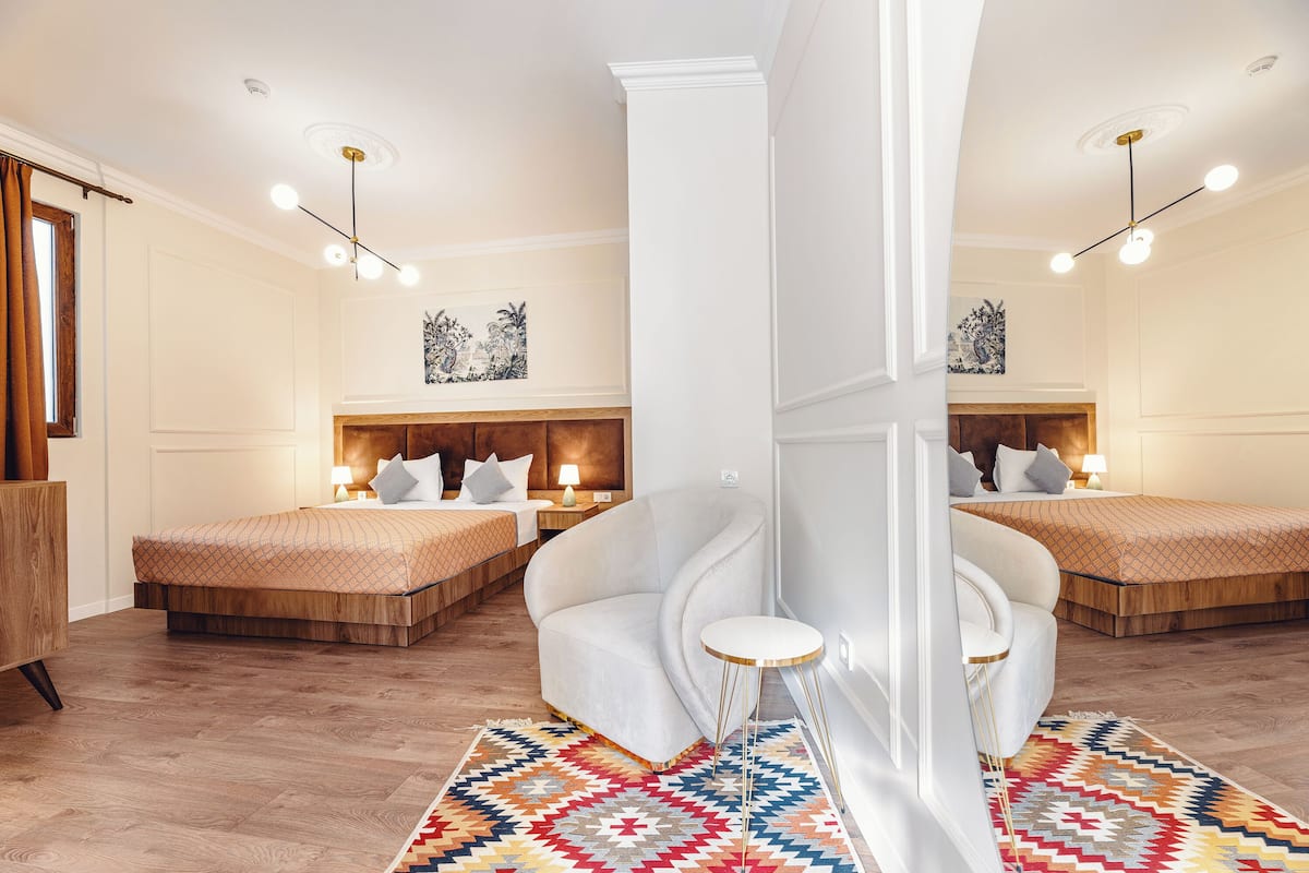 Una habitación de hotel con dos camas, un sillón blanco, una mesita redonda y una alfombra colorida.