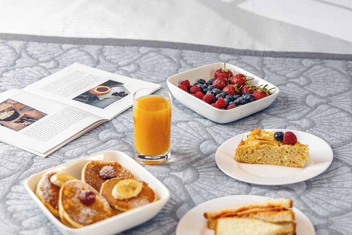 Un desayuno servido en la cama con un libro, un tazón de bayas, panqueques, un vaso de jugo de naranja y una rebanada de pastel.
